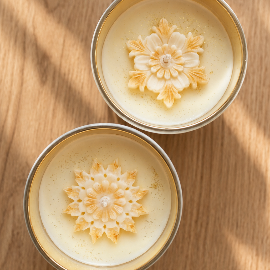 Bovenaanzicht van twee Heaven’s Whisper geurkaarsen met crèmekleurige bloemendecoraties en gouden accenten in houders met goudkleurige binnenkant.