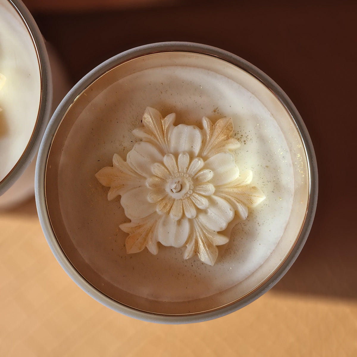 Bovenaanzicht van twee Heaven’s Whisper geurkaarsen met crèmekleurige bloemendecoraties en gouden accenten in houders met goudkleurige binnenkant.